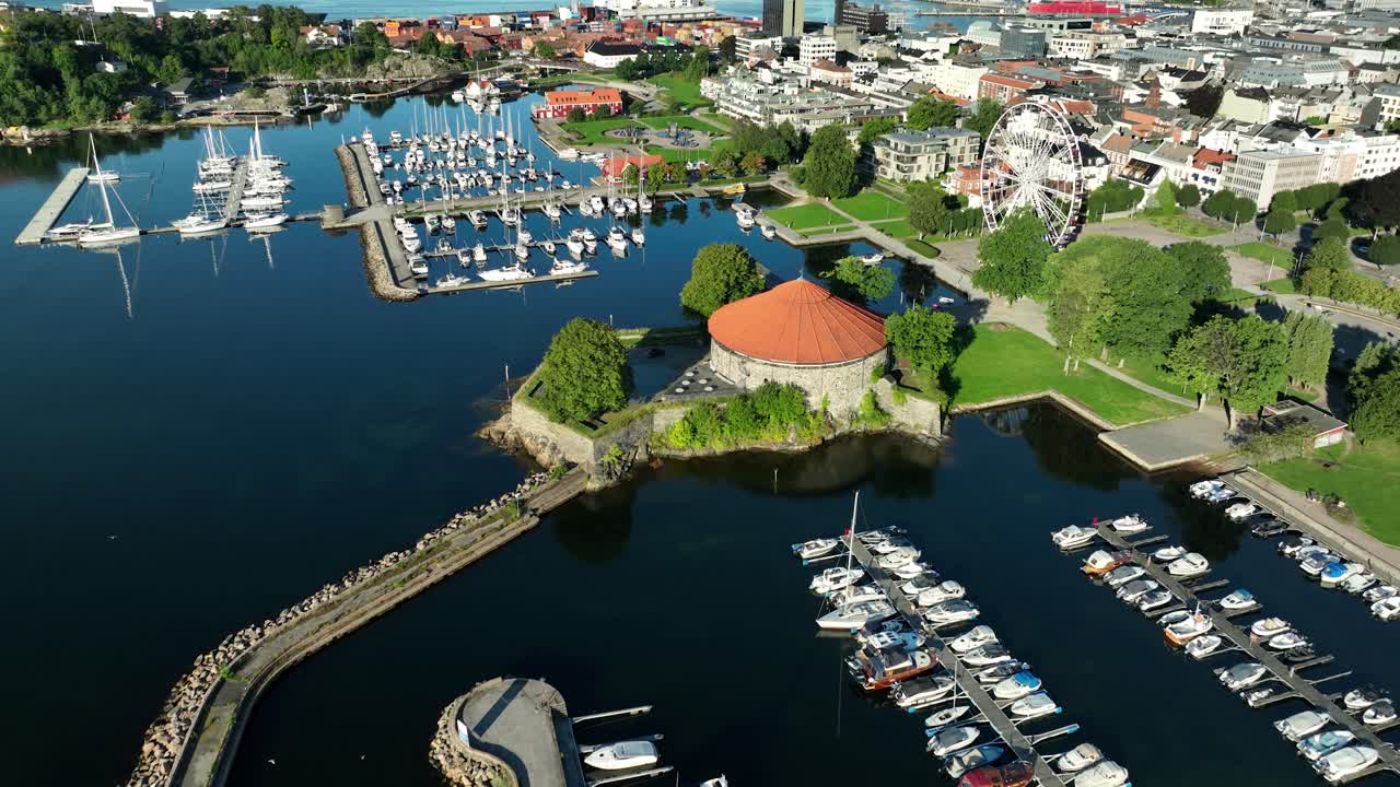 Kristiansand marina filled with sailboats and yachts sits next to a peaceful waterfront. Lush greenery surrounds the area while clear skies reflect off the calm water, creating a picturesque view