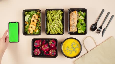 food delivery top view, take away meals in disposable containers. lunch boxes with cooked vegetarian dishes, using phone with chroma green screen. healthy diet. catering service