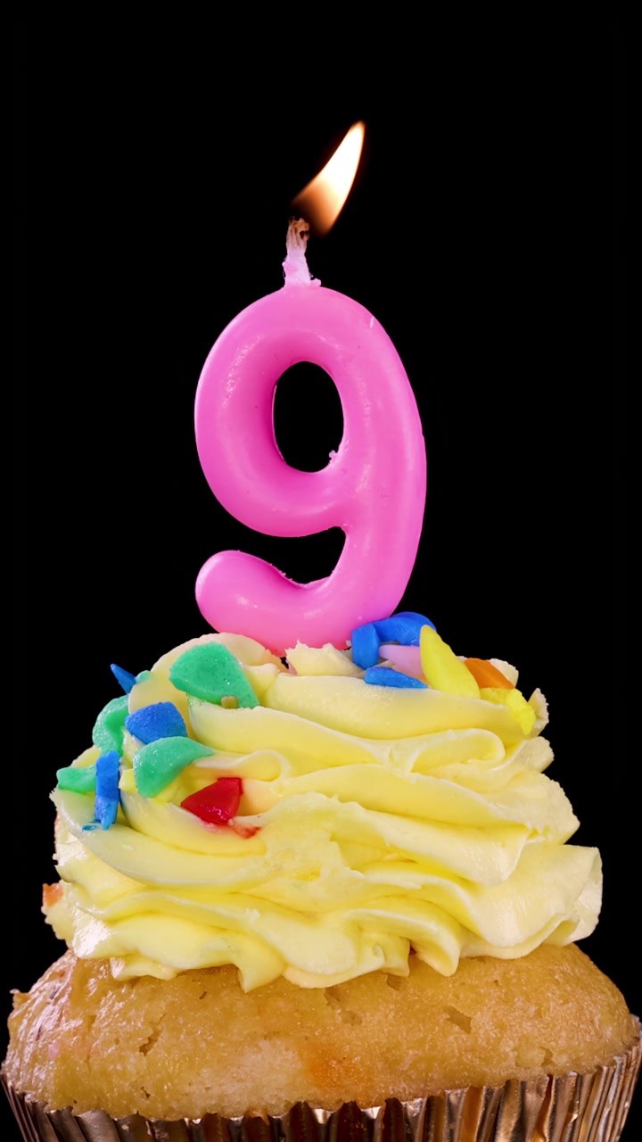 Una candela rosa numero nove brucia sopra un cupcake glassato con zuccherini colorati, su uno sfondo nero con illuminazione brillante e uniforme e senza movimento della telecamera.