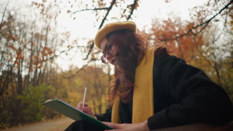 side view of a lady seated sketching in her book, deeply focused against a backdrop of autumn trees and colorful leaves in a tranquil outdoor setting