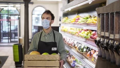 retrato de un hombre guapo con una máscara facial y un delantal avanzando en una tienda de alimentos con una caja de frutas en el interior