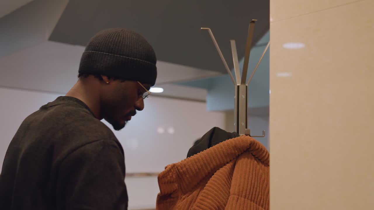 Man hangs coat on lobby rack, knit beanie and textured sweater, warm orange jacket placed, soft overhead lighting, candid commuter moment, interior reception atmosphere, brief arrival gesture