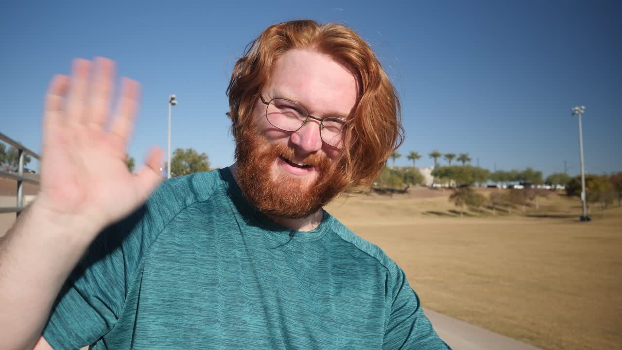 A red haired male smiling and waving at the camera in the park in a close up.