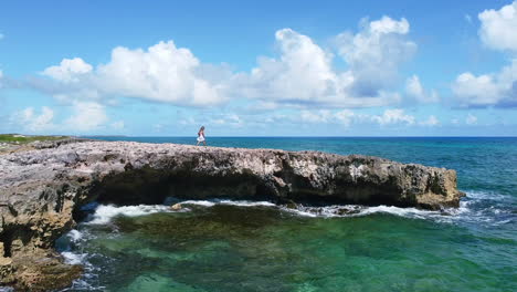 static aerial of young tourist in white dress walking on large rock with waves crashing on coastline in cozumel mexico on a sunny summer day