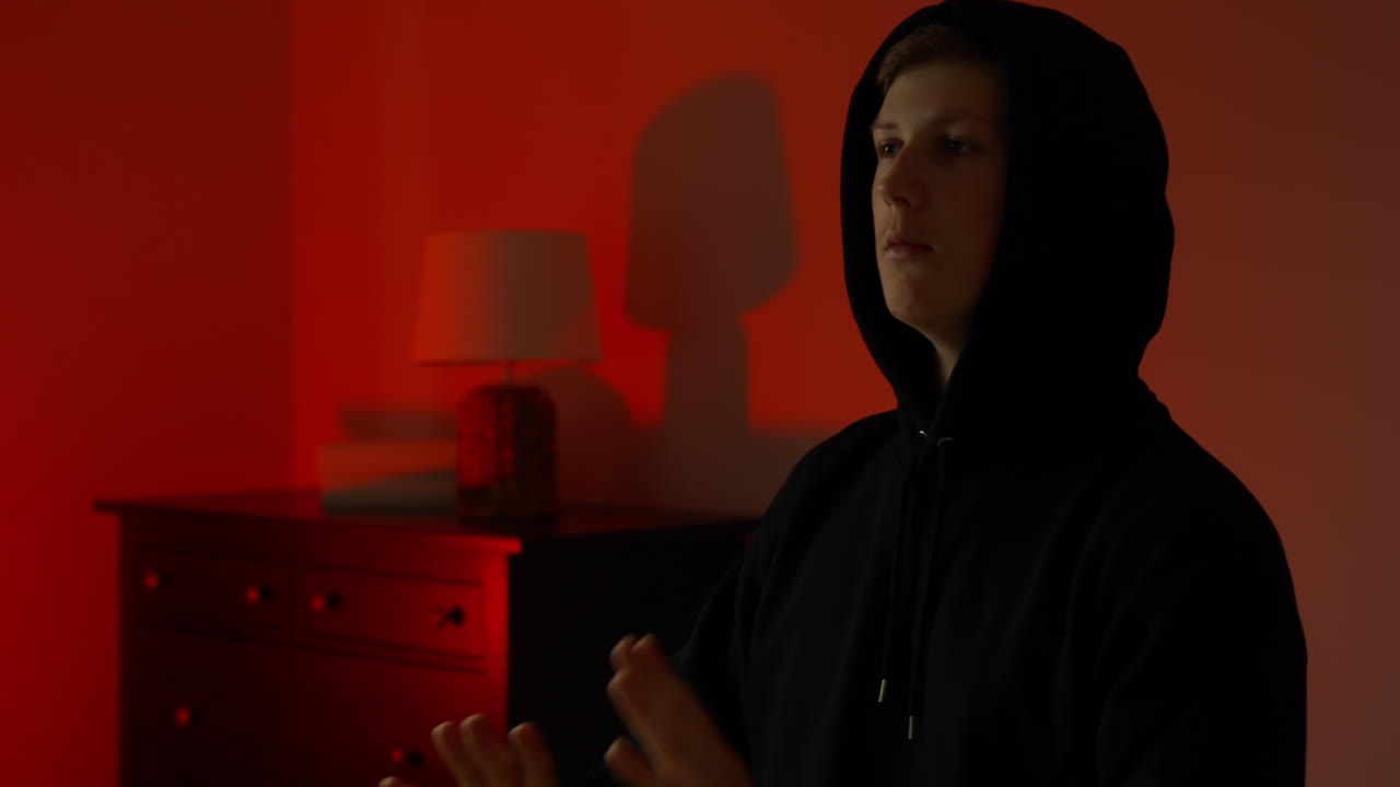 Hooded young man stands inside dark house interior, hands raised in gesture of confusion or surrender, harsh red and blue alarm light emphasizes emotional stress and uncertainty during crime