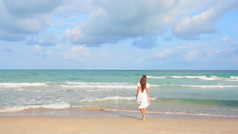 asian woman running barefoot on the beach toward the sea in summer wearing a white sundress then she stops and rais hands up, slow-motion backside view