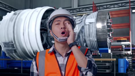 close up of asian male engineer with safety helmet yelling with hand over mouth while standing with airplane engine maintenance conducted, analytics checking the turbine