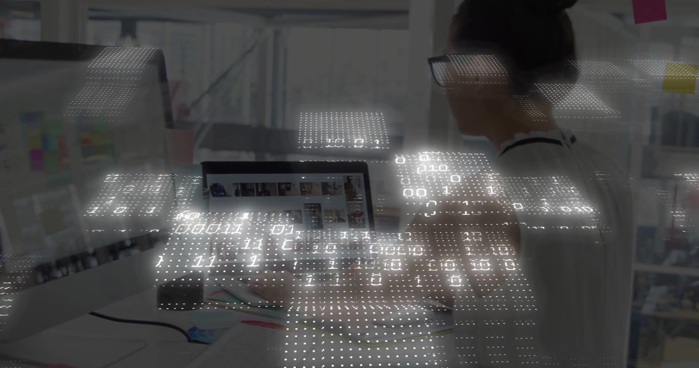 Designerin tippt und klickt, während Laptop und Monitor eine binäre Überlagerung für Datenvisualisierungen anzeigen.