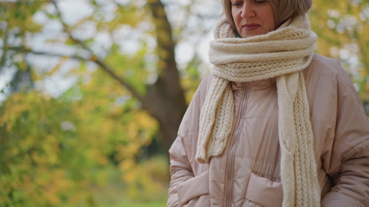 Woman in light jacket and chunky knit scarf walking peacefully with hands in pockets, surrounded by scattered autumn leaves and golden trees, dressed in joggers and sneakers under soft daylight