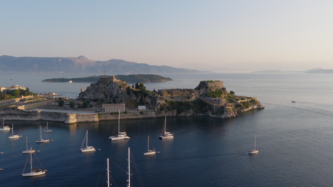 Sunrise light over Corfu Old Fortress and swallows flying above calm sea with anchored sailboats, aerial tracking right