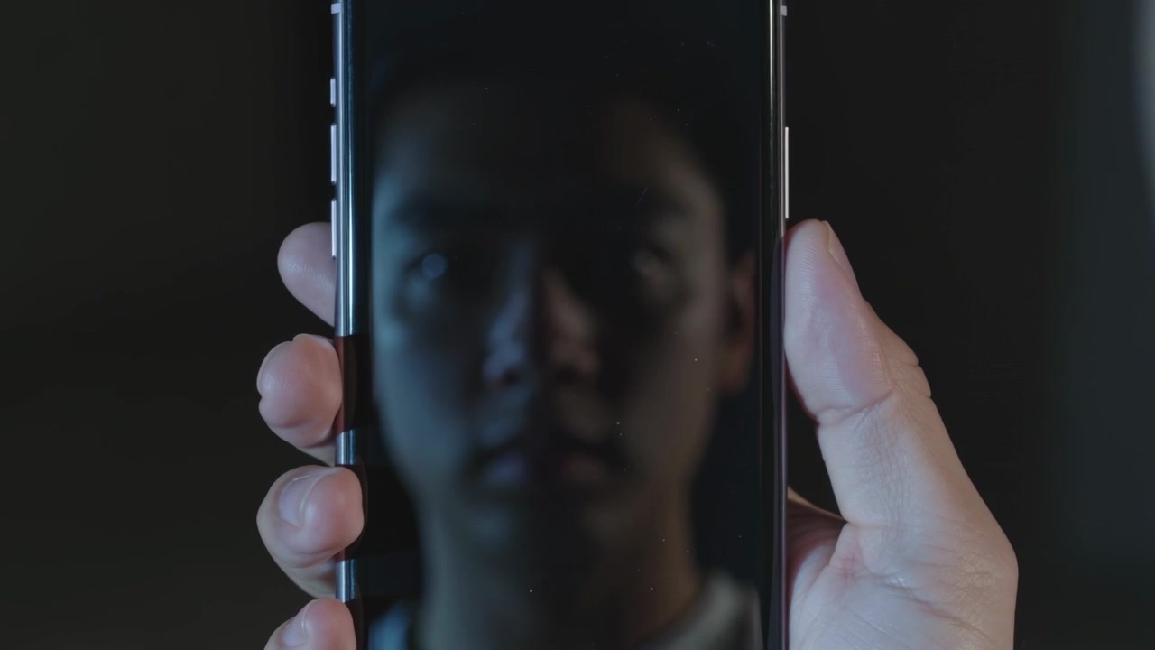 Lifting right hand holding phone reflecting man's face with light collar in dim room checking marks