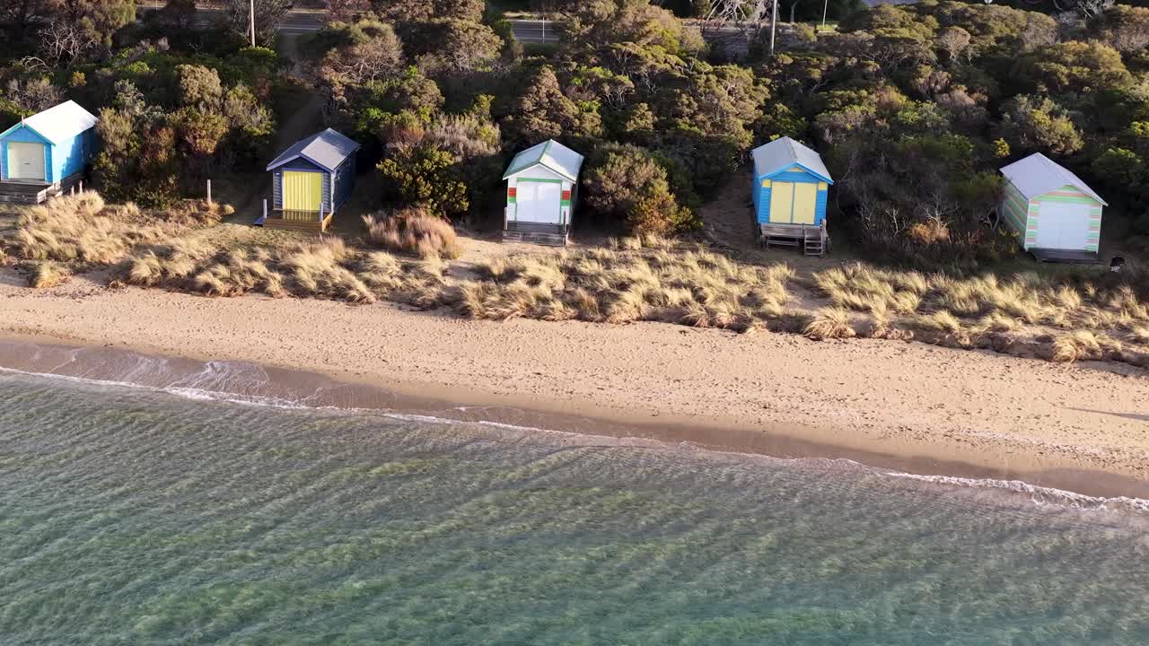 Drone pans above sandy beach and vibrant bathing boxes, morning sunlight, tranquil coastal atmosphere