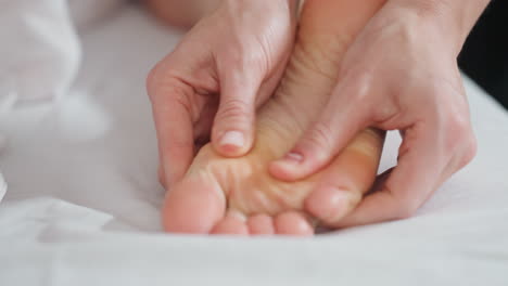 close-up of therapist using both thumbs to gently press client foot and side during massage, focusing on skin texture, hand motion, and therapeutic precision in soft indoor setting with white linens