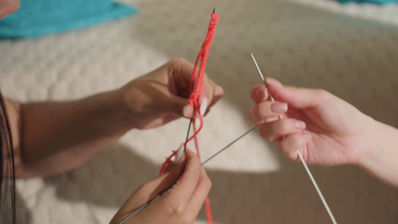 Una pareja caucásica intercambiando puntos de tricotado a la vez en la cama, dos pares de manos pasando agujas y ajustando hilo rojo, momento de aprendizaje cooperativo con trabajo en equipo táctil y comunicación cercana.