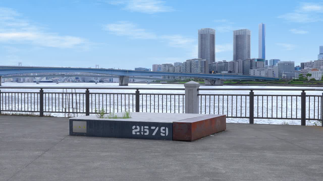 A peaceful shot of a waterfront promenade with a bench, the Harumi Bridge, and the distant Tokyo skyline