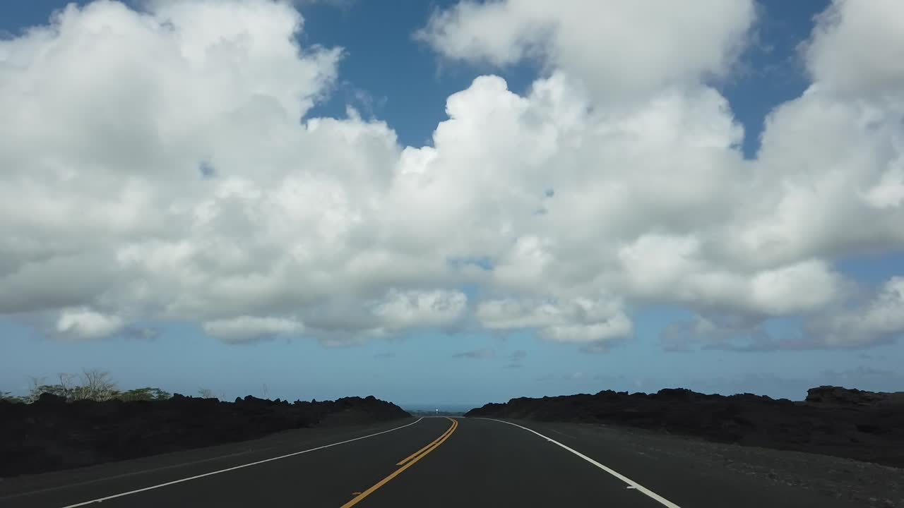 Time lapse of driving along a road passing through a fresh lava field on Hawaii island with the pacific ocean in the background and bright skies with white clouds.