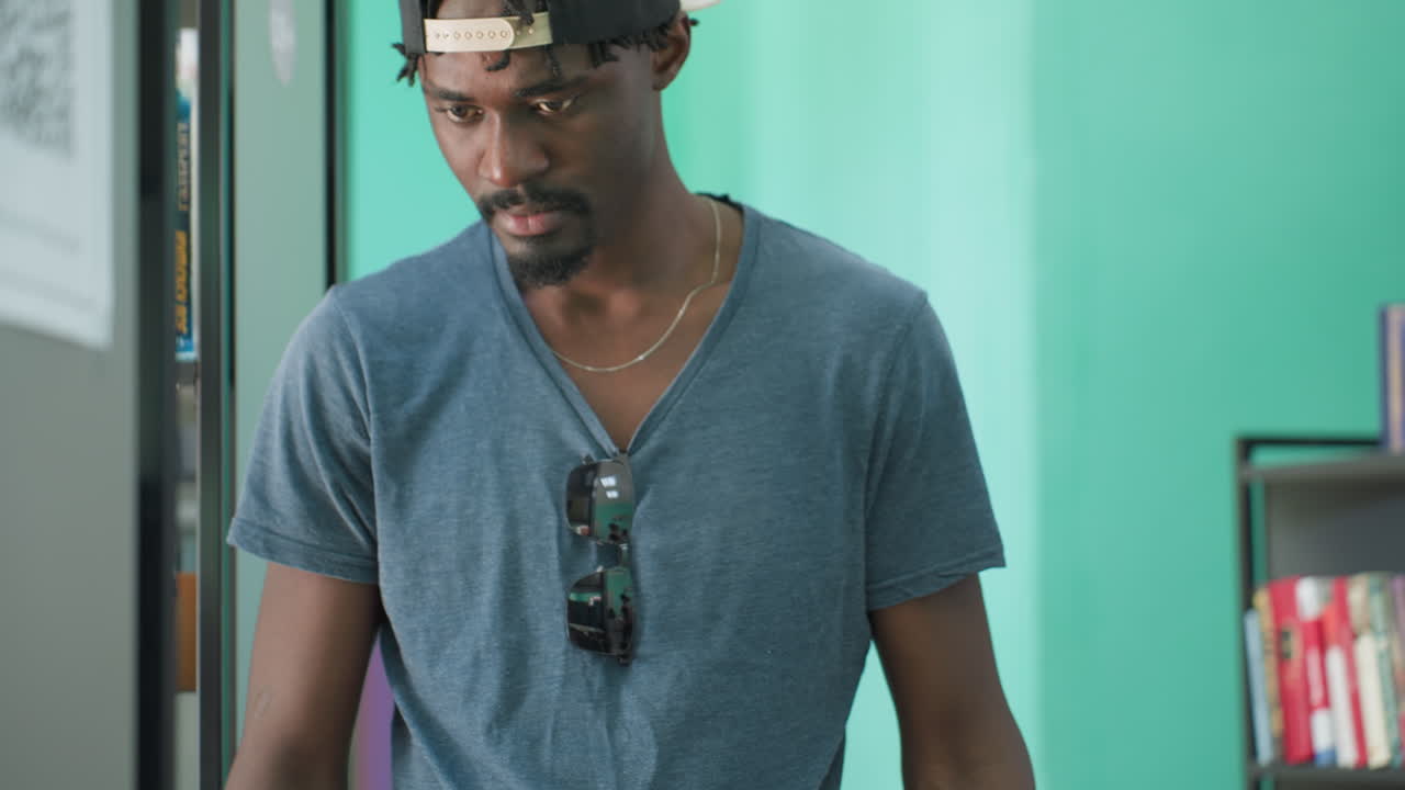 Close up of man in casual t shirt taking books from library shelves, sunglasses hanging on shirt reflecting background scene, against teal wall and visible book spines on nearby shelf