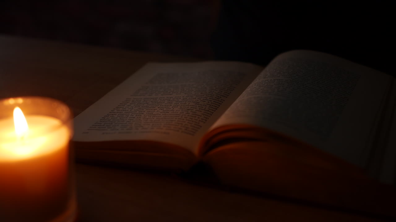A man reading a book by candlelight and putting down his glasses after finishing