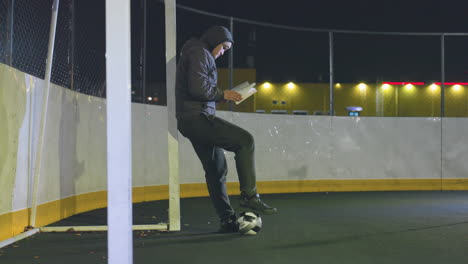 atleta con capucha apoyado contra un poste de meta metálico por la noche leyendo un libro con una pelota de fútbol rodando suavemente bajo los pies, iluminado por las luces vibrantes de la ciudad en un entorno urbano tranquilo y contemplativo