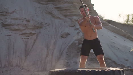 muscle athlete strongman man hits a hammer on a huge wheel in the sandy mountains in slow motion at sunset. the dust from the wheels rises.