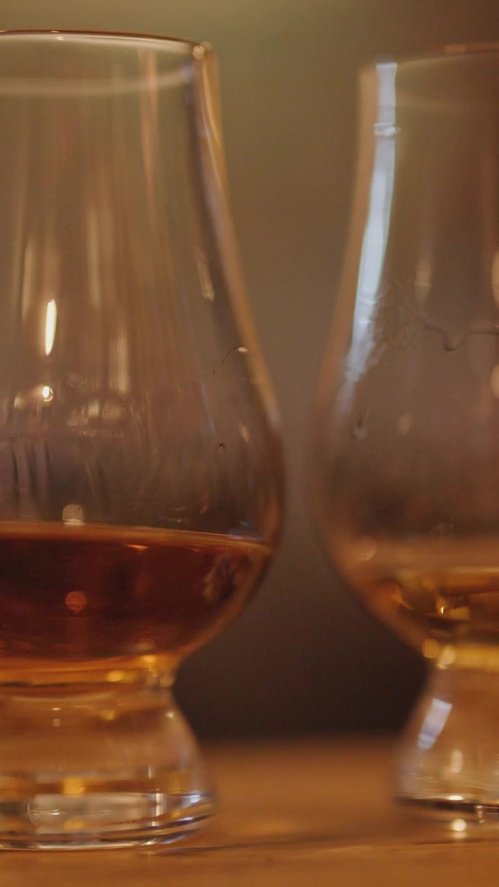 Una mano agita delicatamente un bicchiere di whisky accanto a un altro su un bancone di legno, sotto una calda illuminazione ambientale, con una profondità di campo ridotta e una telecamera stabile.