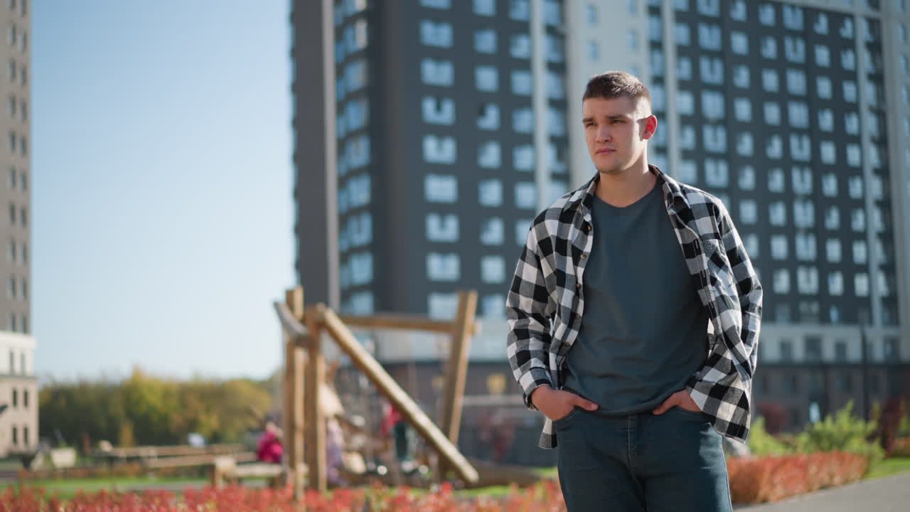 student in walks with hands in pockets while looking around thoughtfully during sunny day in modern residential area with tall buildings and blurred children playing near wooden playground structure
