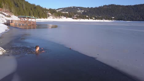 Aerial-views-of-man-sitting-neck-deep-in-frozen-mountain-lake,-drone-rotating