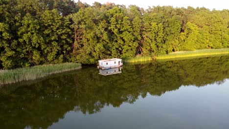 Houseboat-floating-on-a-lake-next-to-reed-during-sunset-in-Brandenburg,-Germany