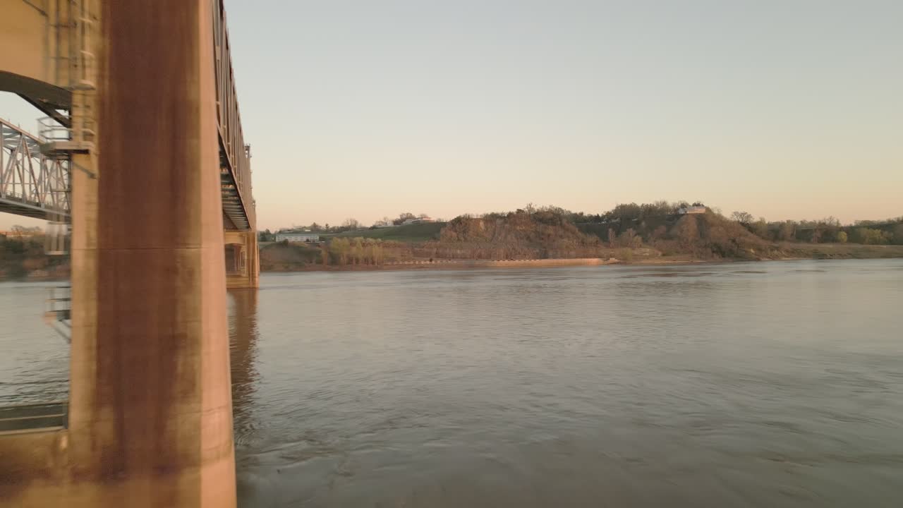 Low drone pass between the twin steel truss bridges over the Mississippi River, warm evening light on the concrete piers and smooth water