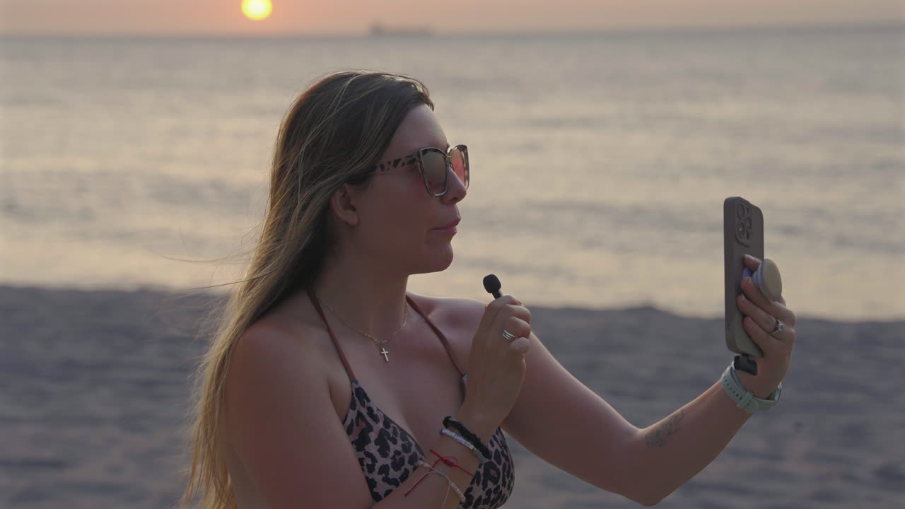 Influencer Bionda Trasmette in Diretta su una Spiaggia al Tramonto Usando un Telefono e un Microfono. - primo piano, ripresa al rallentatore