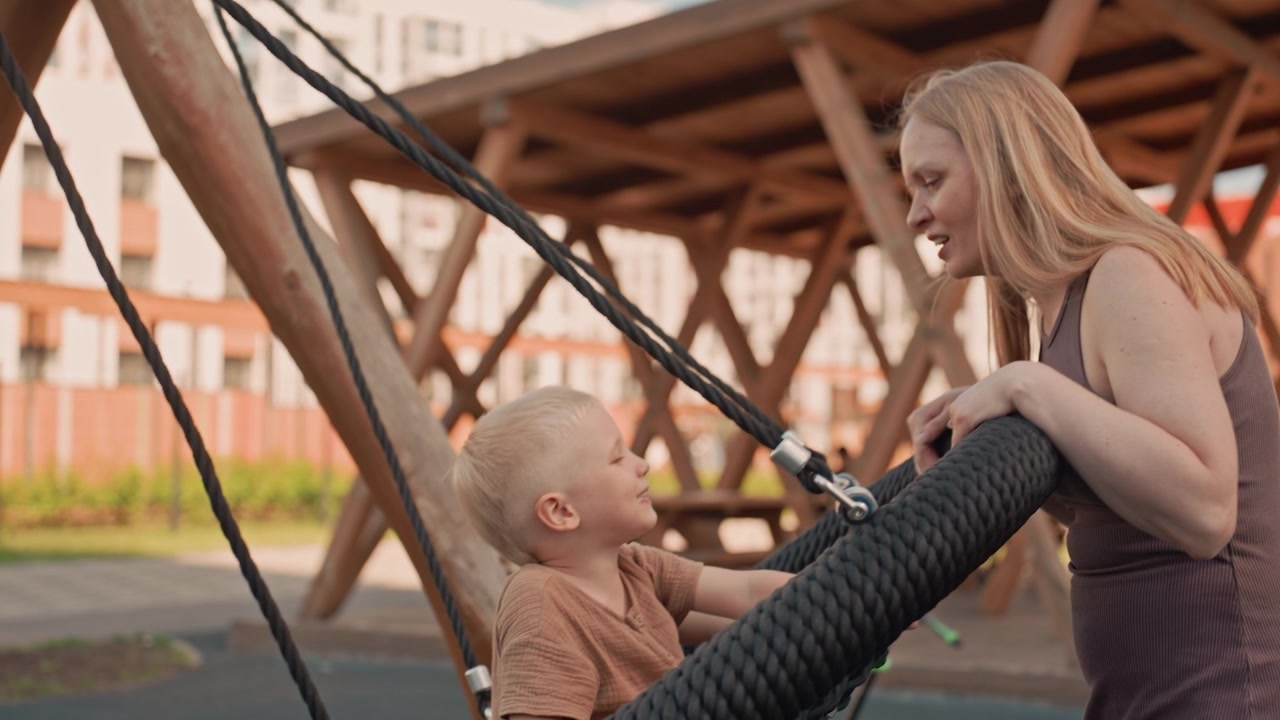 Una donna interagisce con un bambino su un'attrezzatura da parco giochi, spingendo delicatamente un'altalena in un ambiente di parco all'aperto. La scena presenta una luce naturale soffusa ed elementi di sfondo visibili come strutture in legno.