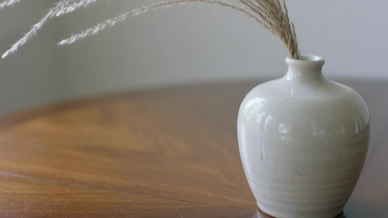 Caméra recadrant et se déplaçant vers la droite, un vase en céramique blanc se déplaçant sur une table en bois, contenant des tiges séchées.