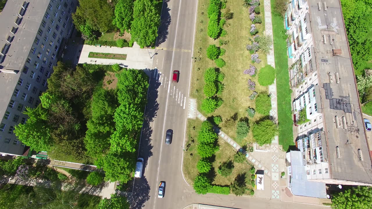 Few cars moving by the empty road through residential area. Multi-storied buildings, cottages and lots of greenery in the outskirts of the town. Top view.