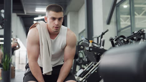 gym trainer sits on weight bench with towel draped over shoulder exhaling in relief after intense chest press workout under bright modern gym lighting showing focused calm expression during recovery