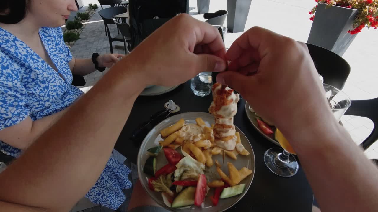 A person enjoying a meal of grilled chicken skewers with fries and fresh vegetables at an outdoor cafe. Ideal for themes related to food, dining, family meals, and casual outdoor eating.