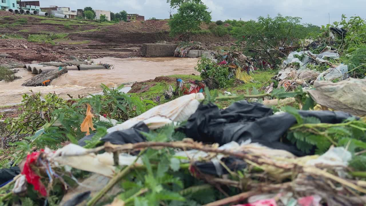 Static shot of a river scene showing environmental degradation. The river itself flows with muddy, sediment-laden water, indicating high levels of suspended solids likely due to erosion or runoff
