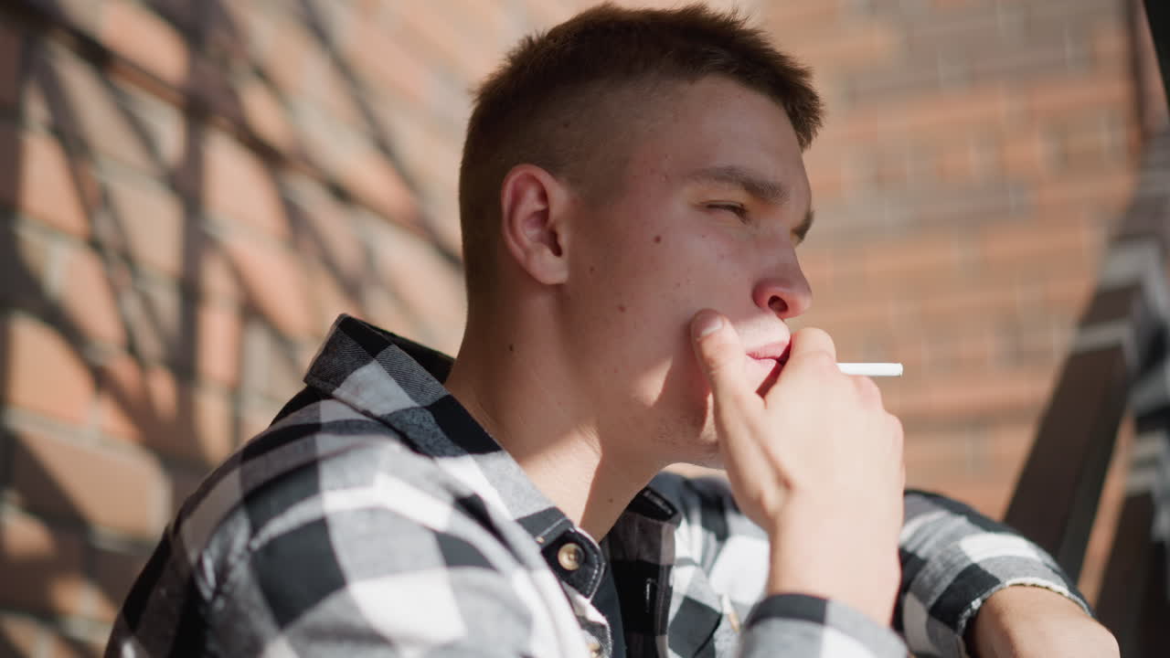side view of man resting on iron railing smoking cigarette gazing thoughtfully into distant wearing check flannel shirt and jeans warm sunlight casts geometric brick and staircase