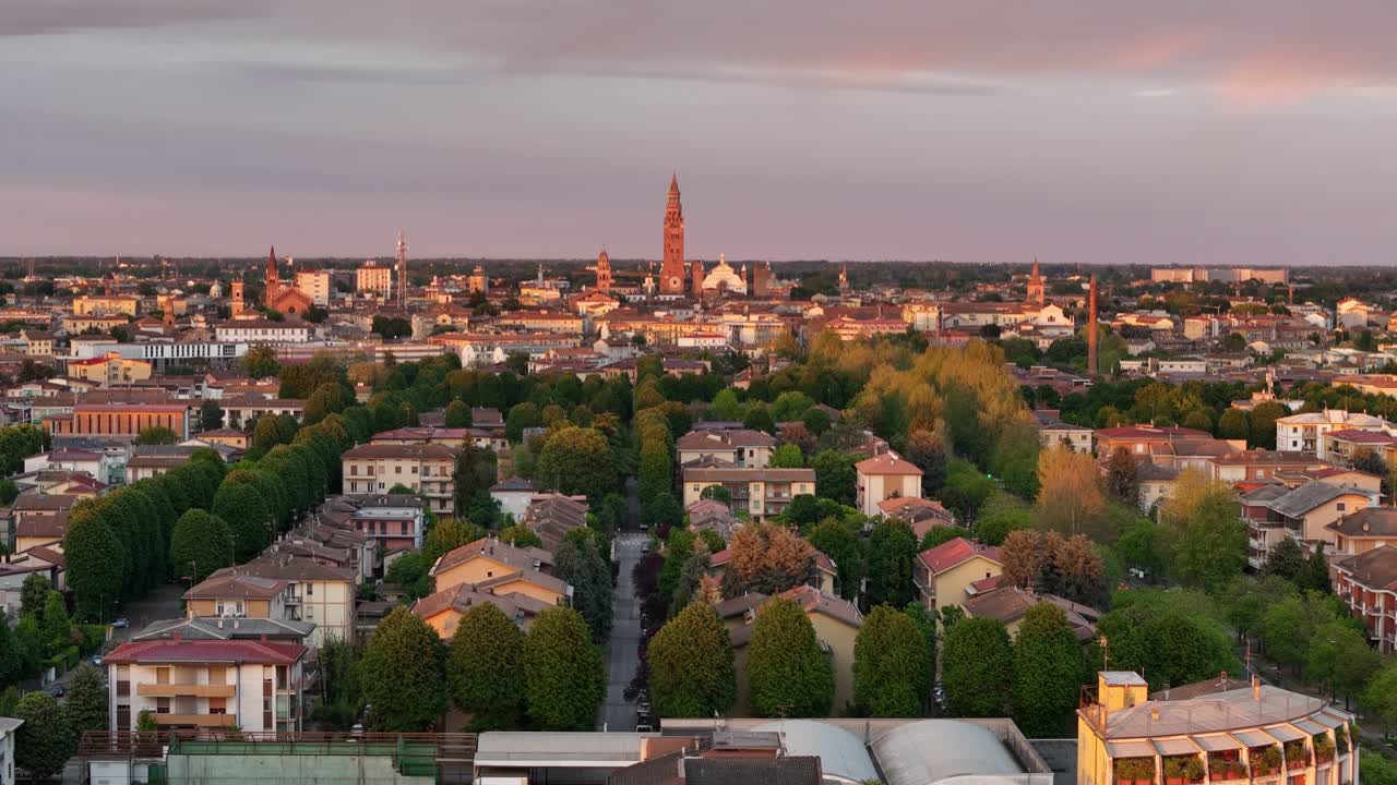 Drone ascends above Cremona, revealing tree-lined avenues, historic rooftops, and iconic landmarks including the Cathedral of Cremona and the towering Torrazzo, bathed in golden sunset light