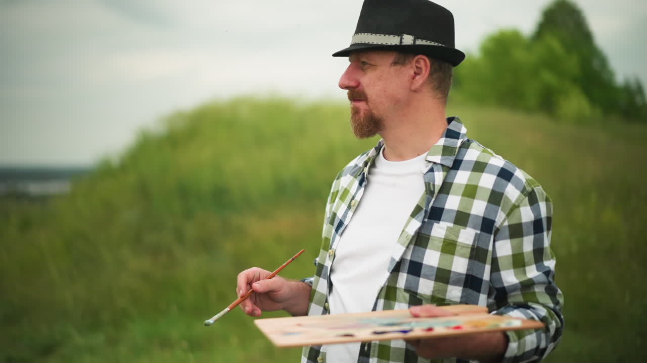 Close-up of a painter wearing a hat and checkered shirt, standing focused in a serene grassland near a lake. He holds a palette and a paintbrush, staring thoughtfully at the landscape