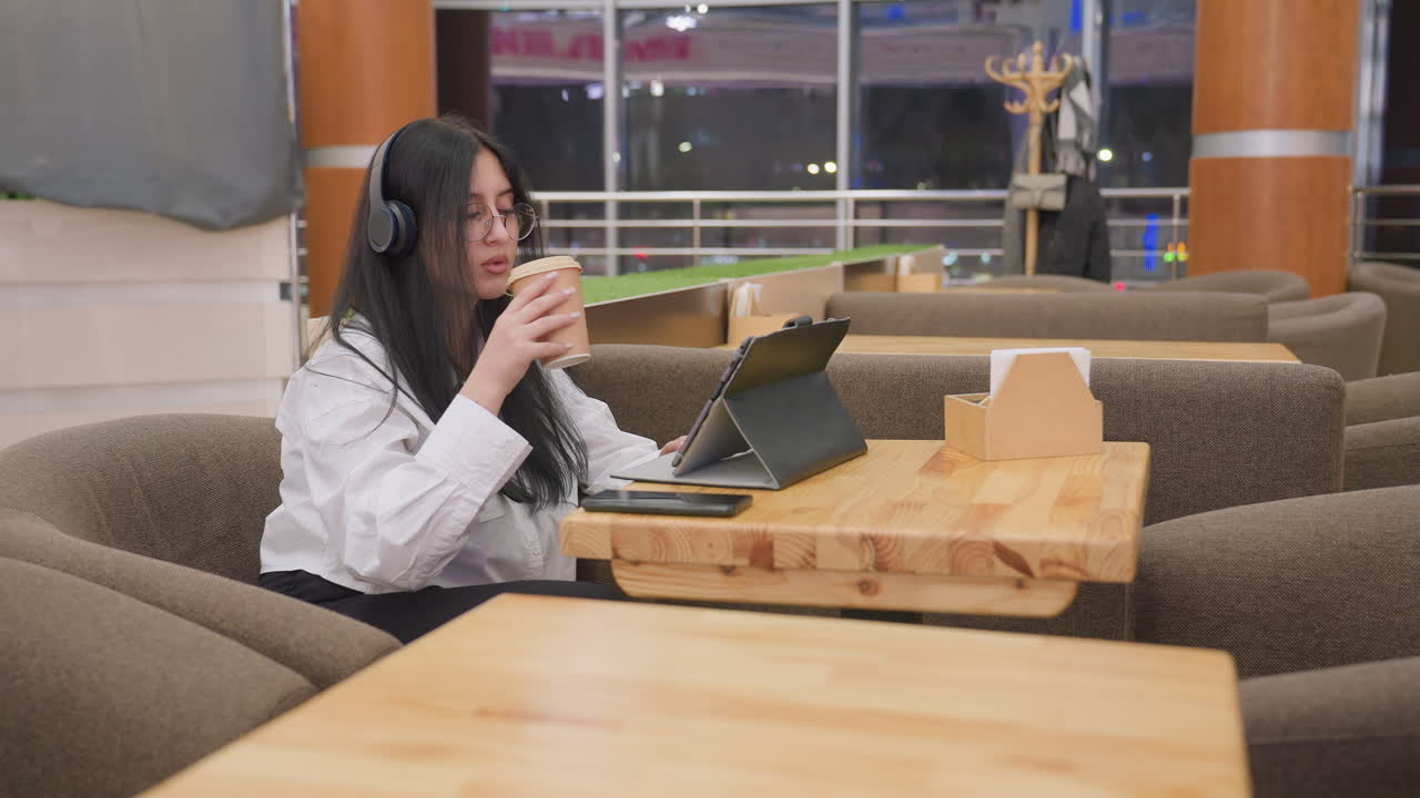 young woman with long dark hair wearing headphones and glasses seated in modern lounge working on tablet with takeaway coffee on table and indoor lighting reflecting warm and calm environment