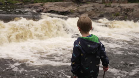 little boy standing at the edge of a river watching the white water rapids