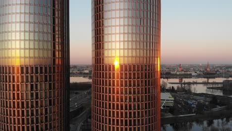 Seitliche-Drohnenaufnahme-Aus-Der-Luft-Von-Zwei-Hochhausbürotürmen-Aus-Glas,-Die-Den-Goldenen-Sonnenuntergang-An-Fenstern-Reflektieren,-Mit-Riga,-Lettland-Stadtbild-Im-Hintergrund-Mit-Klarem-Violettem-Himmel