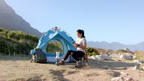 Camping in mountains, woman sitting by tent using smartphone, enjoying nature