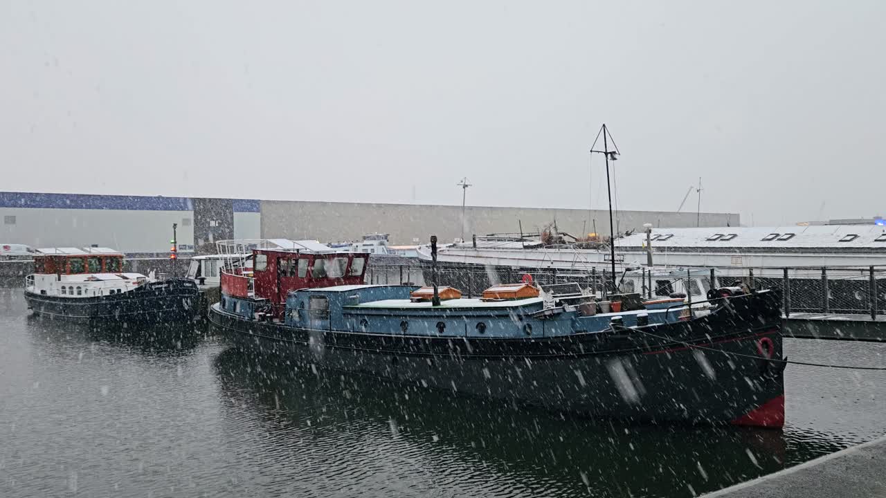 A row of living boats moored in a residential dock during heavy snowfall, with calm water and industrial buildings in the background