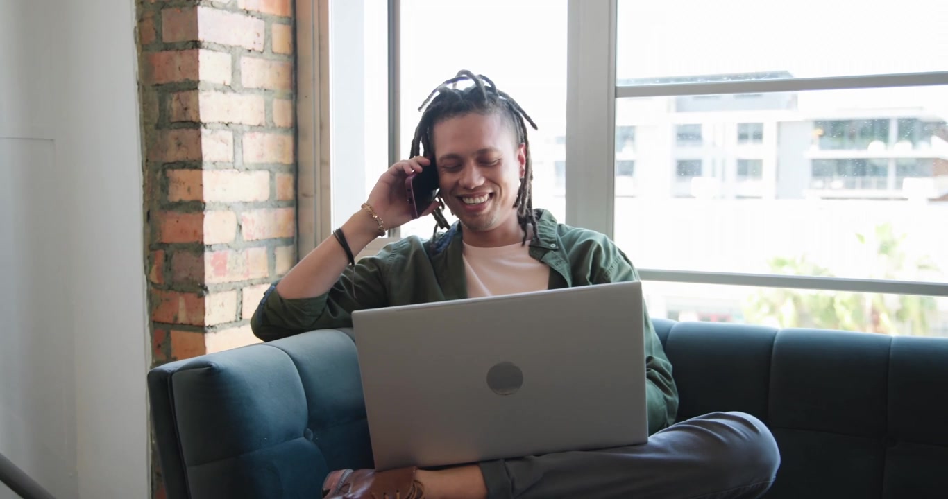 Adult African American man taking incoming call, laughing, typing on laptop, working on blue couch