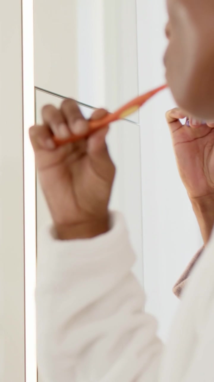 Video vertical: Mujer trayendo un cepillo naranja, cepillándose los dientes frente al espejo en bata, aseándose, con espacio para texto.