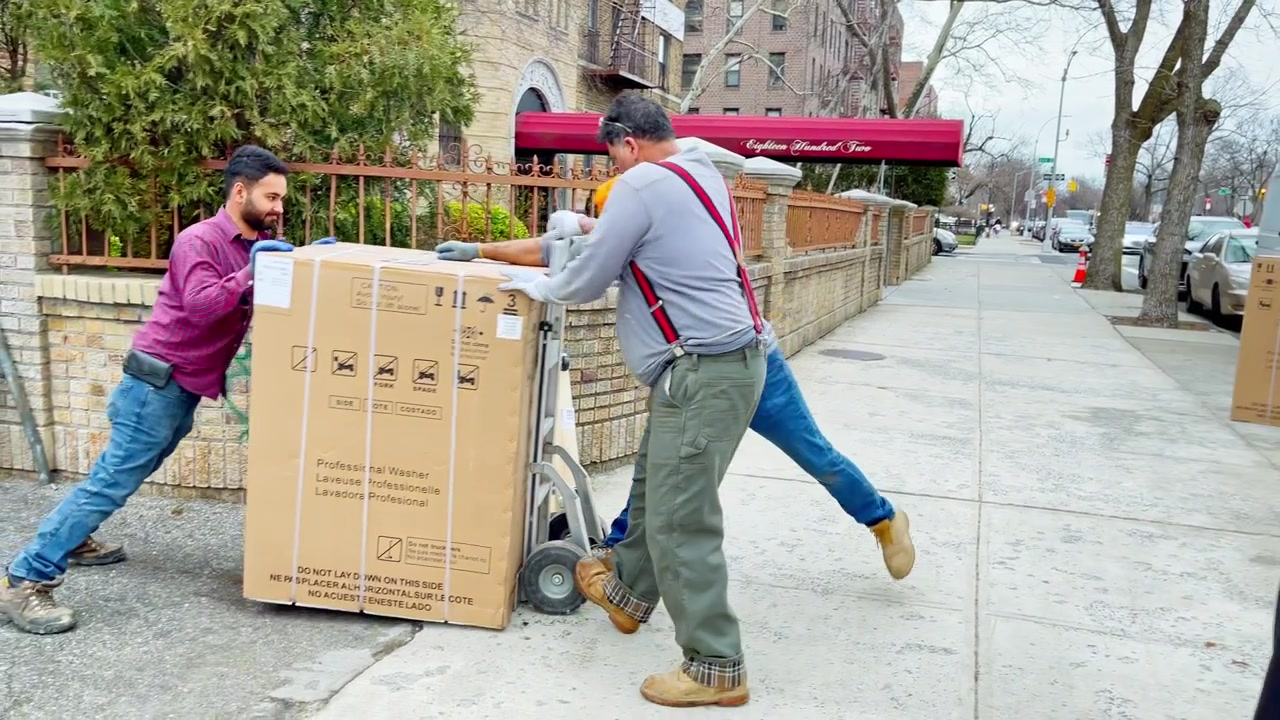 New York, USA, 8 May 2025 : Delivery workers unloading large packaged appliance on sidewalk. Group of delivery men handling a large boxed appliance on a dolly along a city sidewalk in daytime.