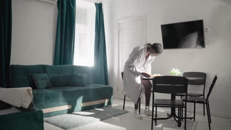 man in white robe standing and placing cup of tea on table in modern living space. sunlight streams through window, highlighting cozy, clean, and inviting atmosphere in well-lit room