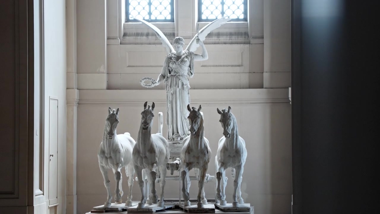 Witte quadriga sculptuur van een gevleugelde overwinningsfiguur en vier paarden staat in een hoge binnenhal, omlijst door zachte raamgloed.