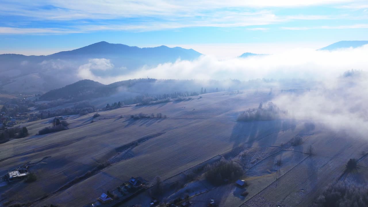 Aerial hyperlapse video of rolling hills, fog drifting over frosty fields, and scattered rural villages under a bright blue sky.